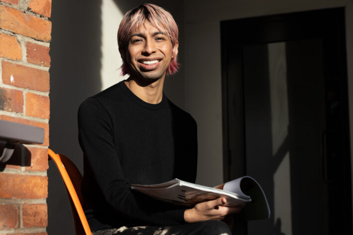A smiling person with pink-tinted hair holds an open magazine in warm sunlight