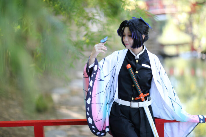 A cosplayer in a butterfly-winged anime costume holds a small blue butterfly on a red bridge