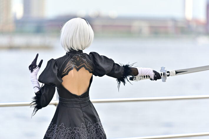 A cosplayer in a black dress and white wig holds a sword, seen from behind by the water