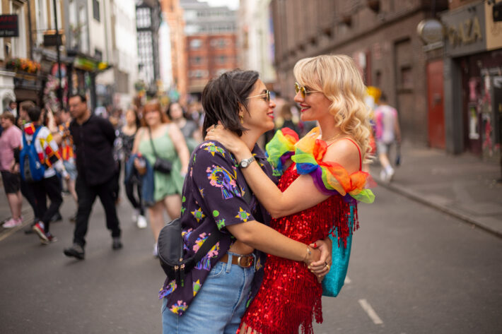 Two people in colorful outfits embrace and smile on a city street during a Pride celebration
