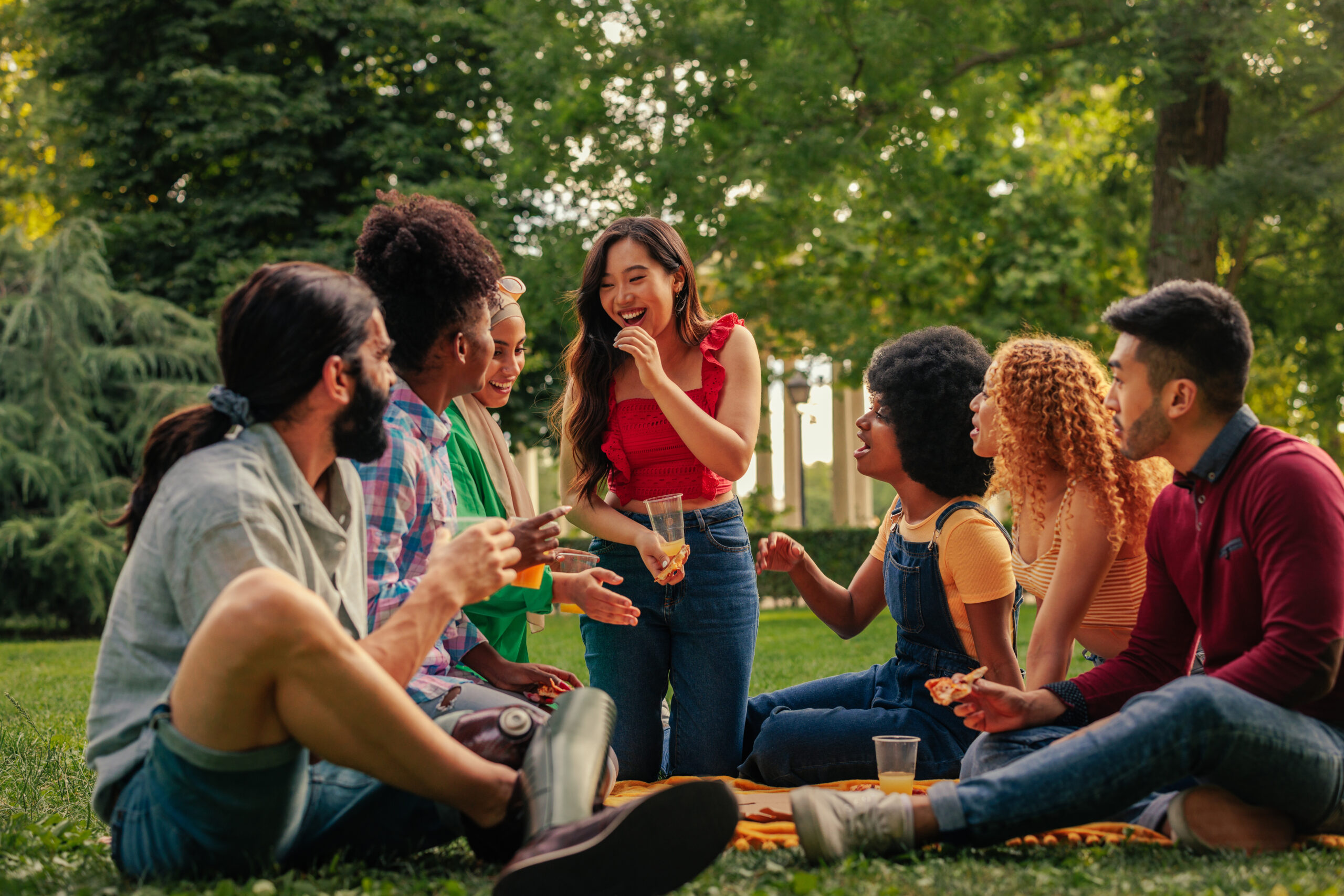 A diverse group of friends sit on the grass in a park, talking and sharing food